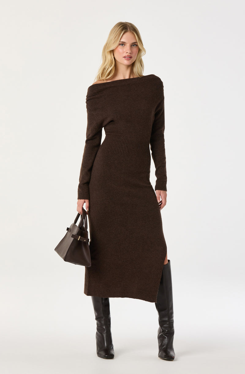 Woman in a long brown knit dress with a slit, holding a gray handbag and wearing black knee-high boots.