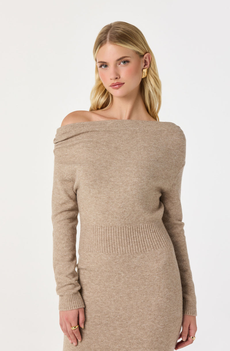 A woman is wearing a beige, off-the-shoulder knit sweater dress with long sleeves. The sweater has a wide neckline that drapes over her shoulders. The dress features a ribbed waistband detail. She is accessorized with gold earrings and rings.