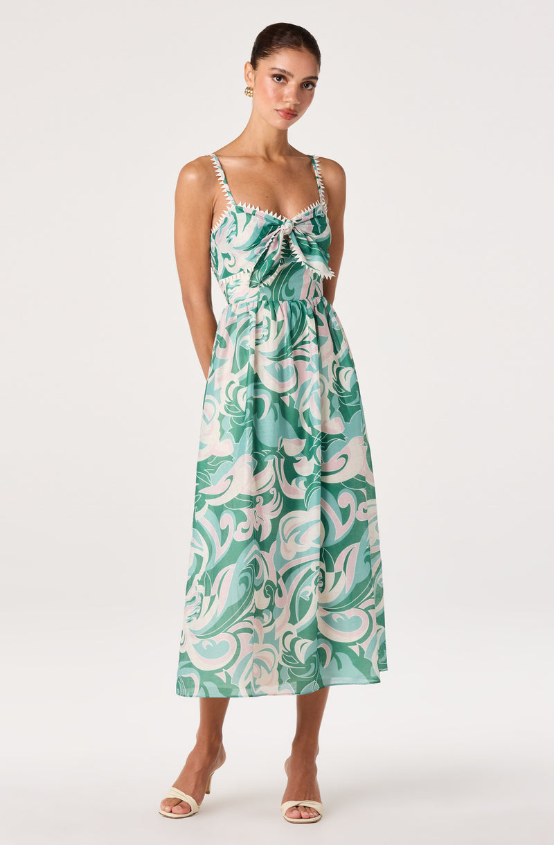 Woman in a sleeveless, green and white patterned dress with a bow at the front, ankle-length.