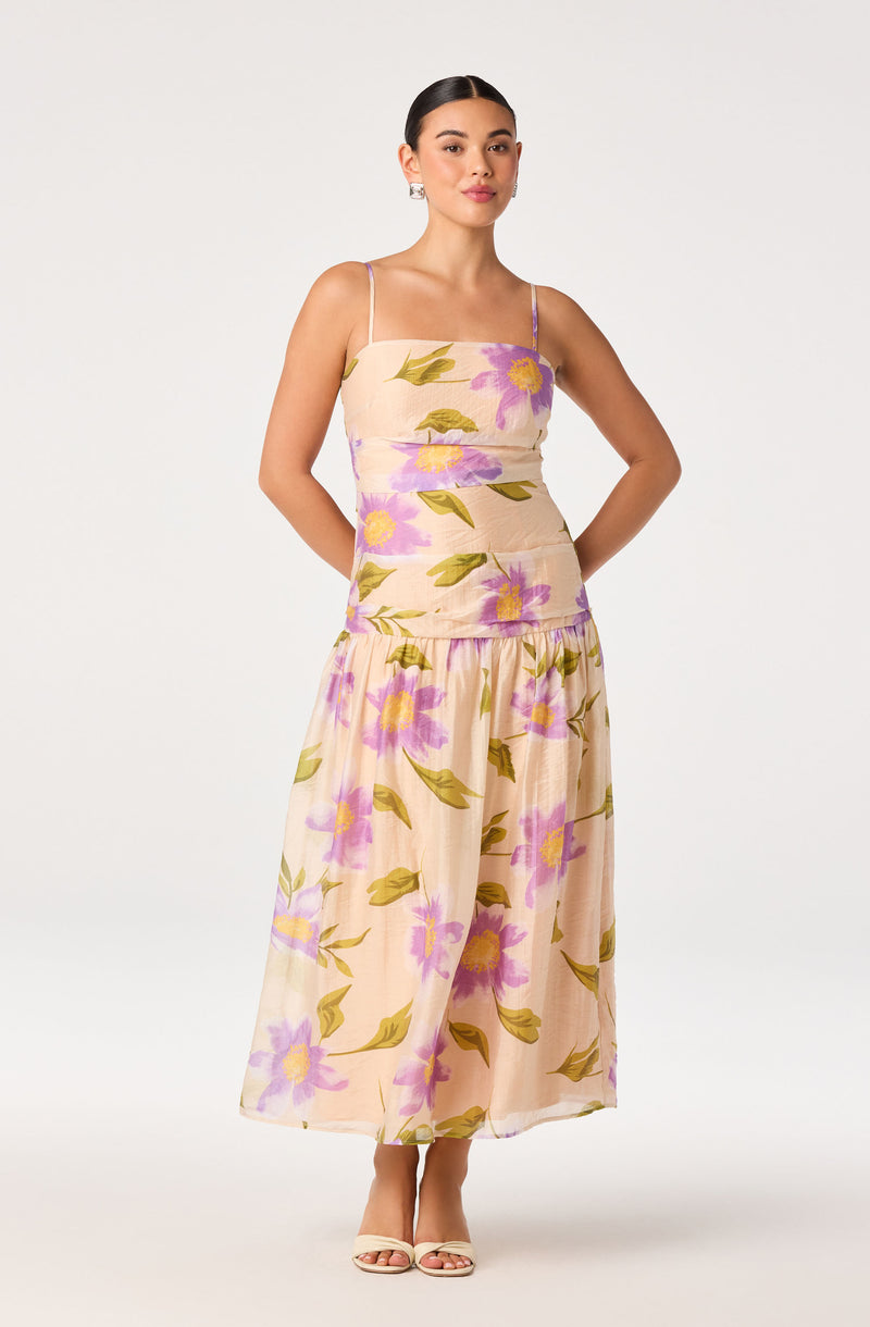 Woman in a light beige floral dress with purple and yellow flowers and green leaves, spaghetti straps, and a layered skirt.
