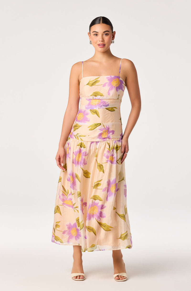 Woman in floral print beige dress with purple and yellow flowers, spaghetti straps, and open-toe heels.