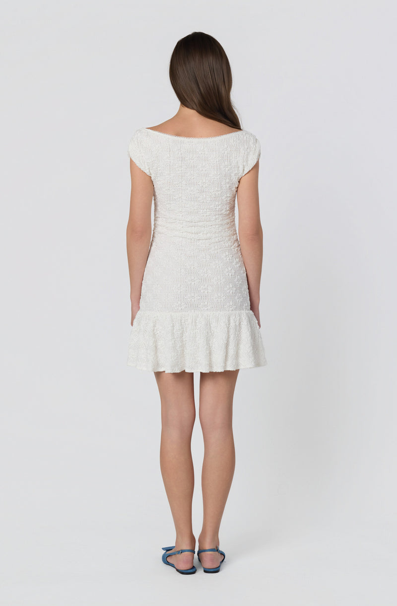 Woman in ivory textured dress with a ruffled hem and cap sleeves, wearing blue sandals.