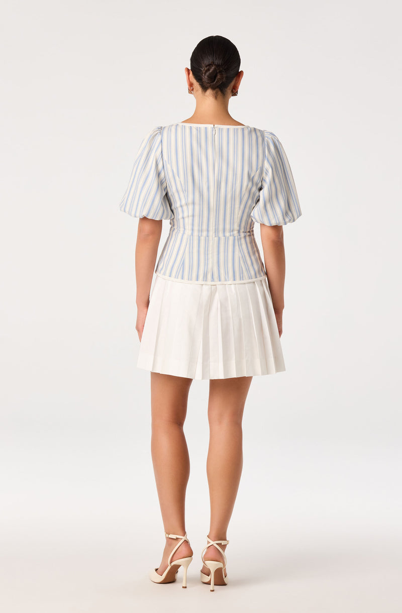Back view of woman wearing a blue-striped puff sleeve top and a white pleated skirt, with beige high heels.