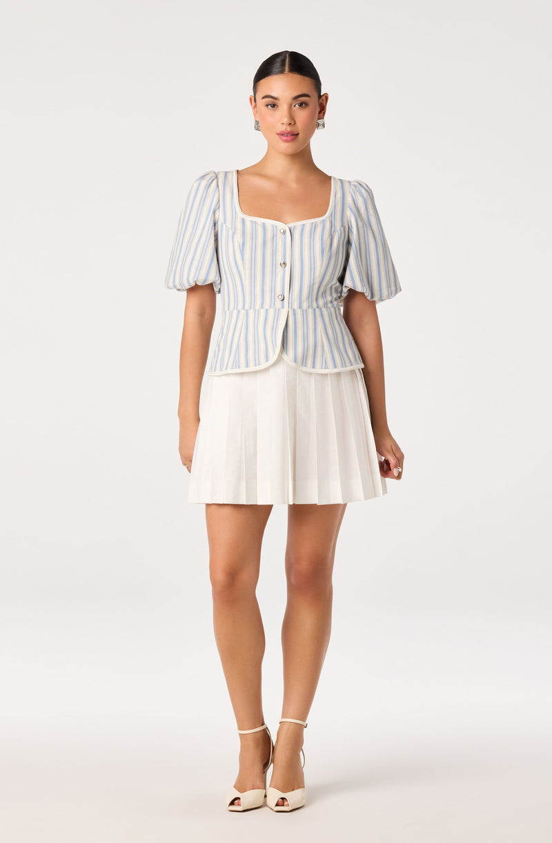 Woman in a light blue and white striped puff-sleeve top with buttons, paired with a white pleated skirt.