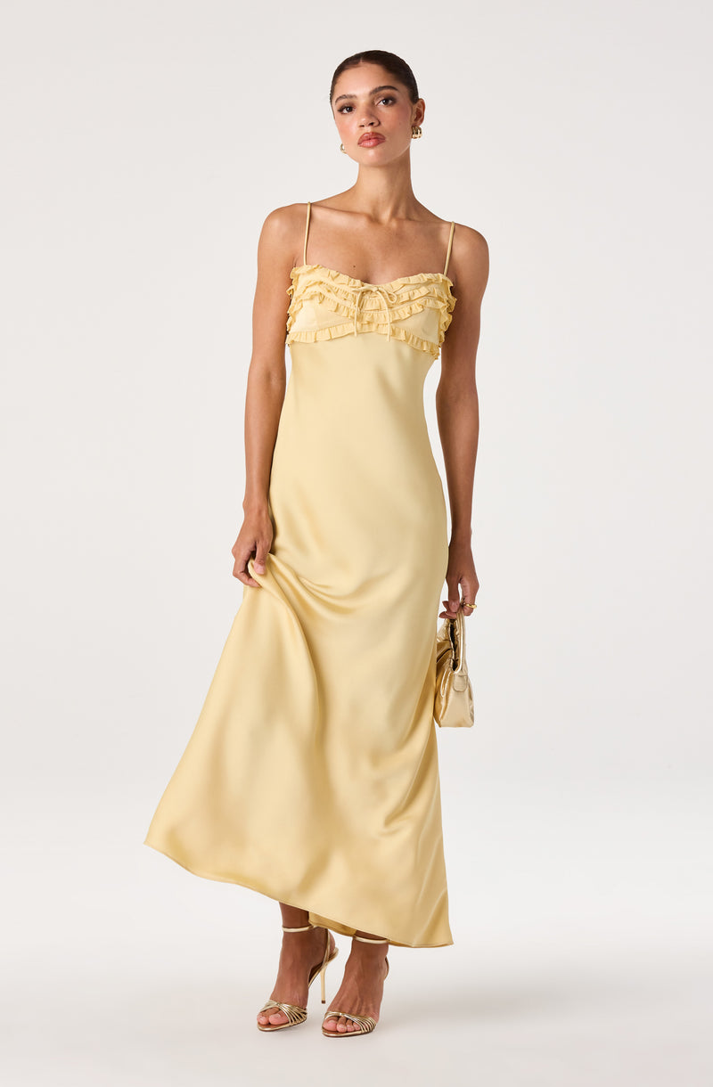 Woman wearing a yellow satin dress with spaghetti straps and ruffled detail on the chest, paired with gold high-heeled sandals and a small matching handbag.
