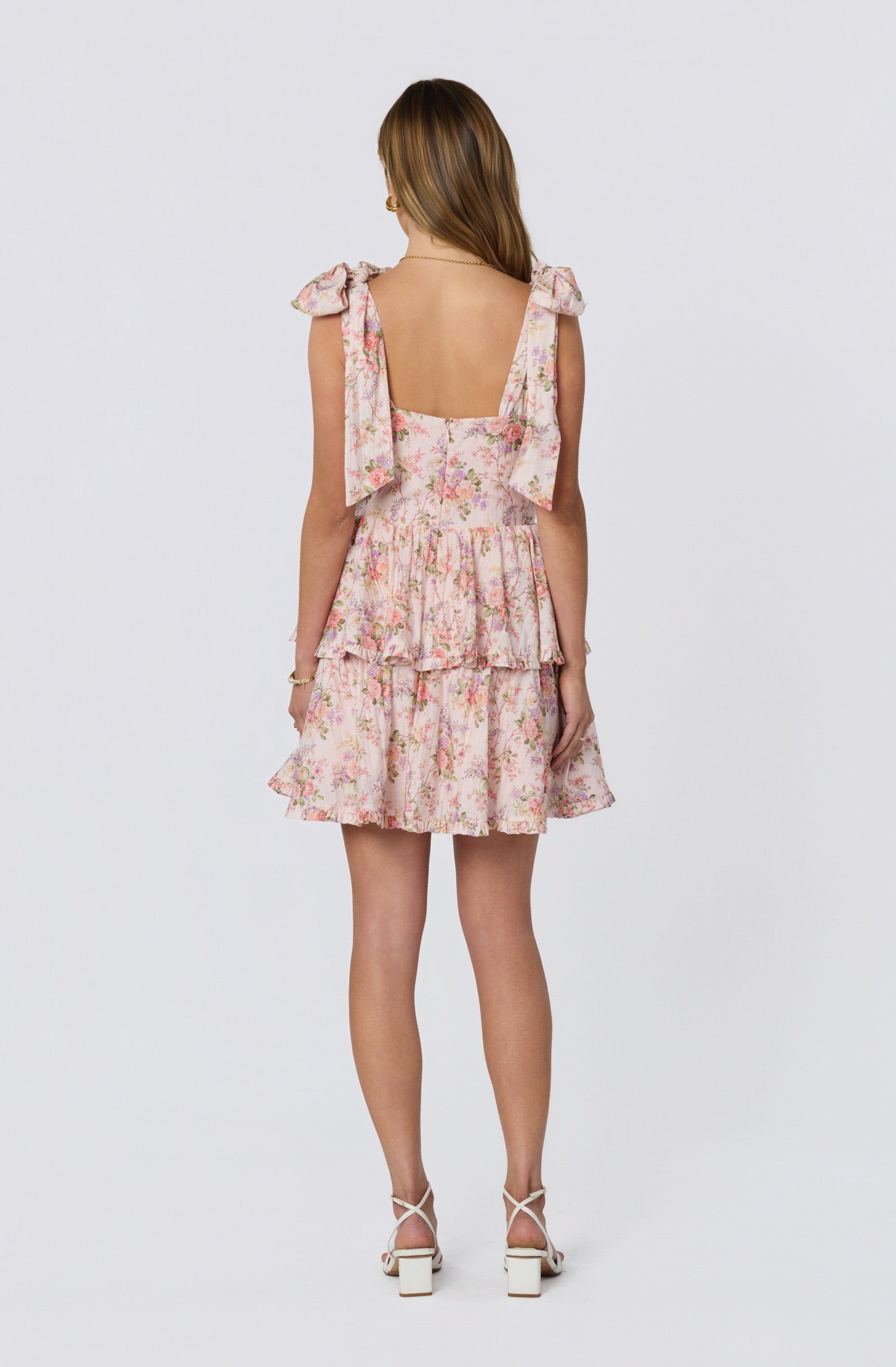 Back view of a woman wearing a pink floral tiered dress with shoulder bows and white high-heeled sandals.