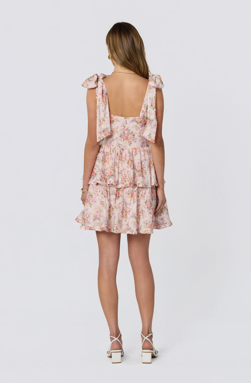 Back view of a woman wearing a pink floral tiered dress with shoulder bows and white high-heeled sandals.