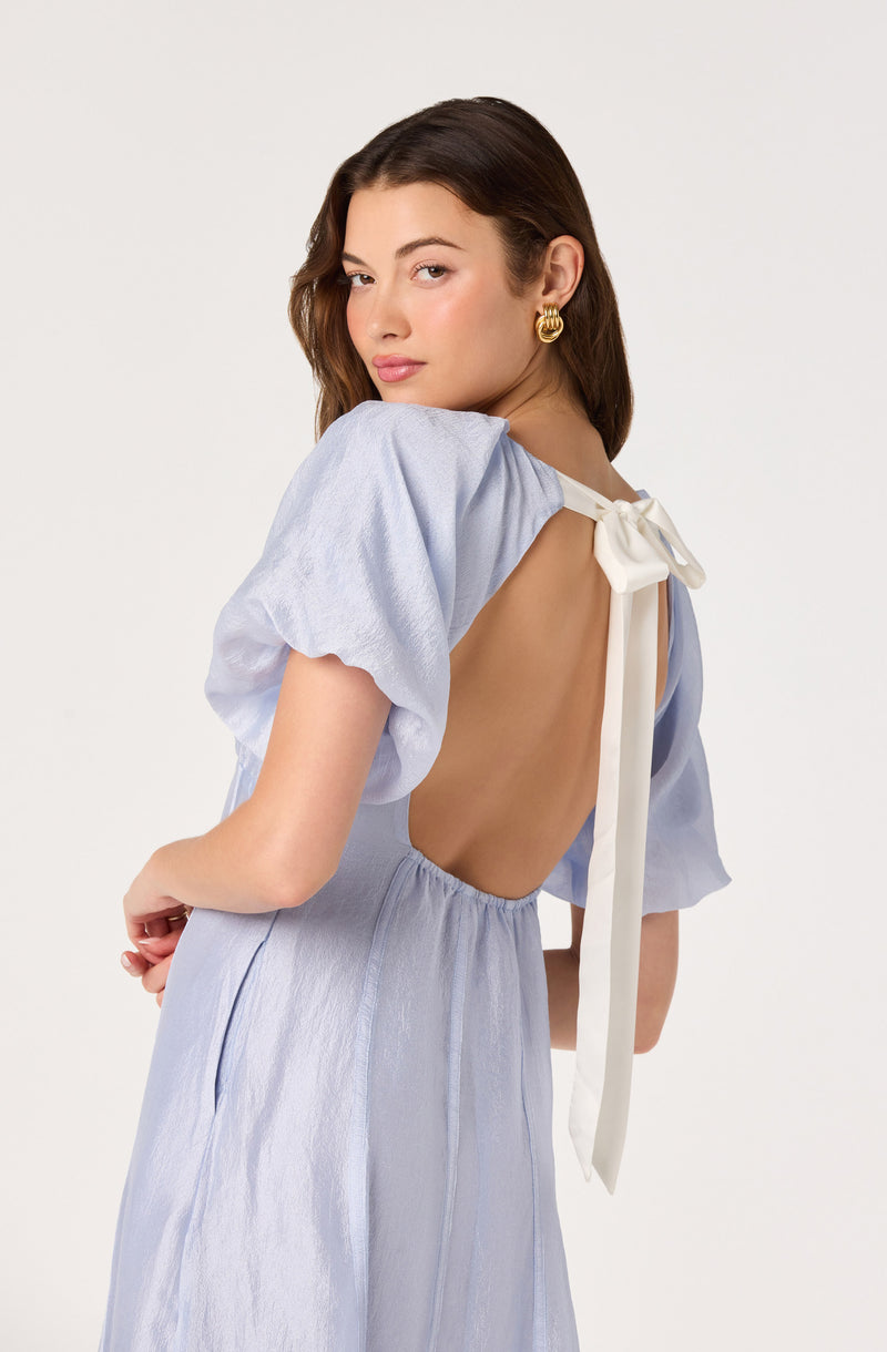 Girl wearing a light blue dress with puffed sleeves and a backless design with a white bow.