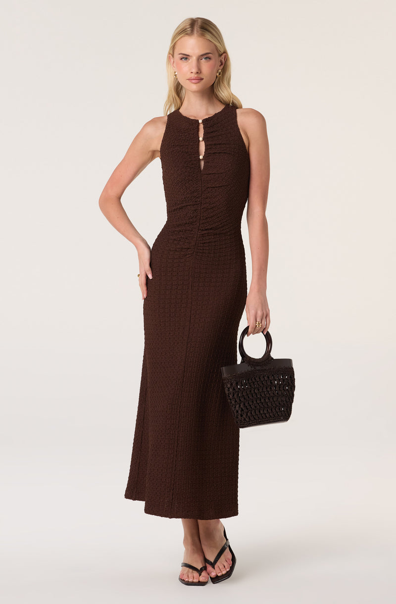 Woman in a sleeveless, textured brown maxi dress with cutout details at the neckline, holding a black woven bag.