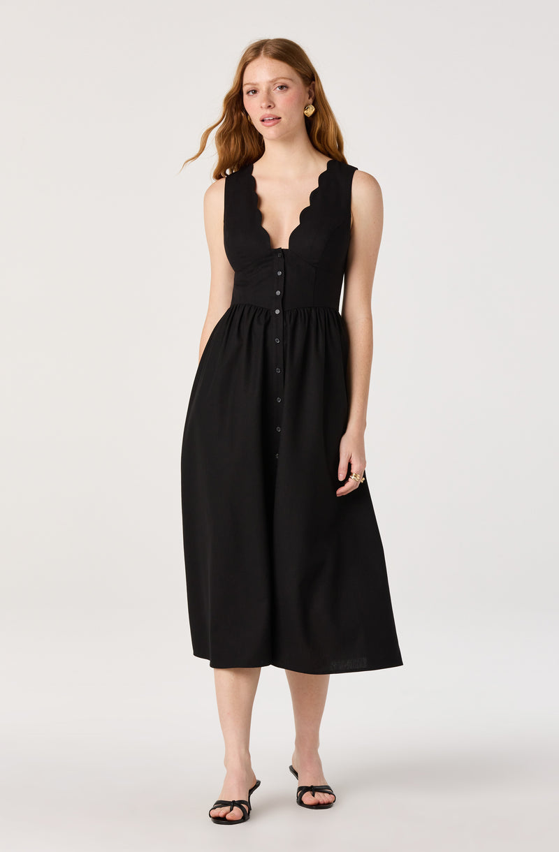 Woman in a black sleeveless midi dress with scalloped neckline and button details, wearing black sandals.