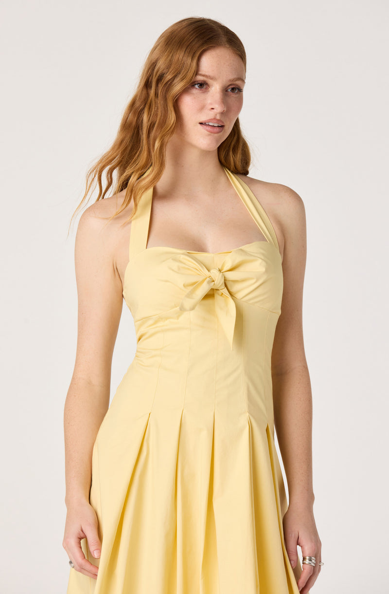 Woman wearing a light yellow dress with a bow at the chest and pleated skirt.