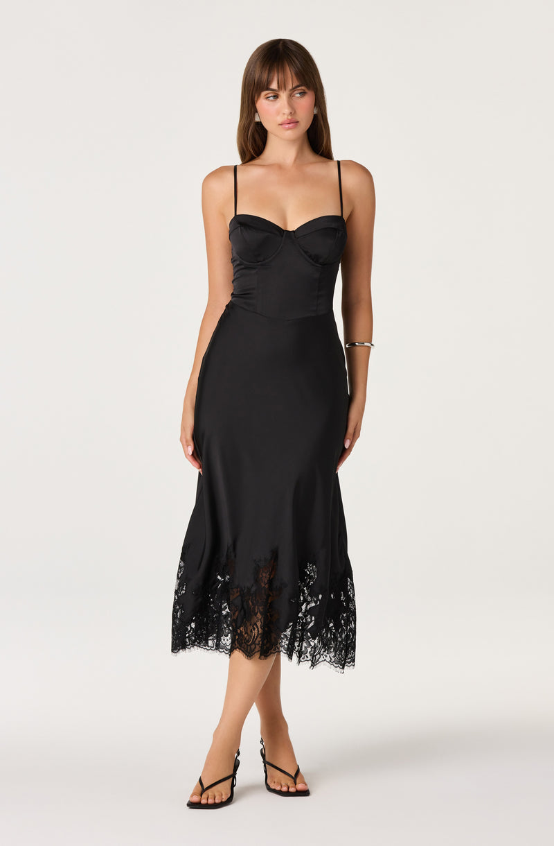 A woman modeling a black spaghetti-strap midi dress with a sweetheart neckline, fitted corset-style bodice, and a lace-trimmed hem, photographed against a plain light gray studio background.