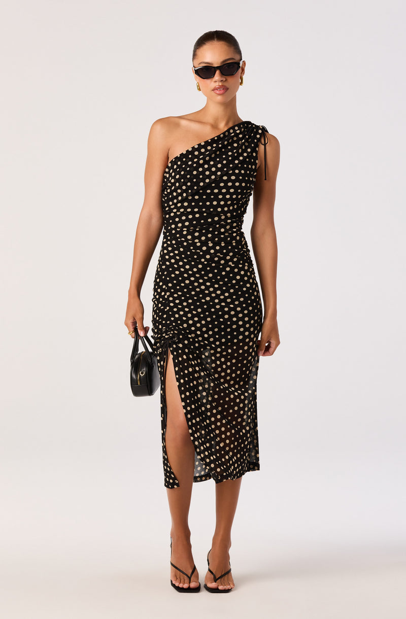 Woman in a black and beige polka dot one-shoulder dress with a slit, holding a black handbag.