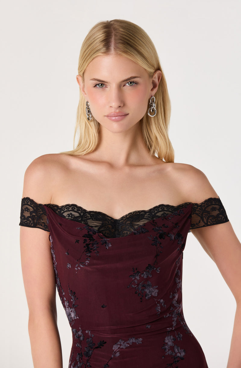 Woman in off-the-shoulder maroon dress with black lace details and floral pattern, wearing dangling earrings.