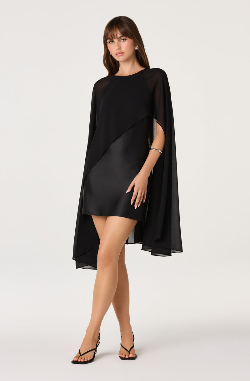 Woman in a black asymmetrical dress with sheer cape-like sleeves and high heels.