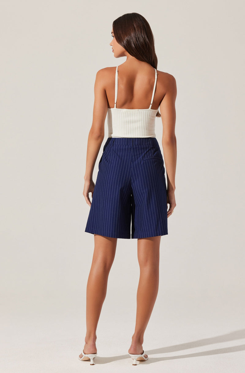 A woman is wearing a white sleeveless top with thin spaghetti straps and a fitted, ribbed texture. She is also wearing navy blue shorts with white pinstripes, which are loose-fitting and reach above the knees. She has on white high-heeled shoes.