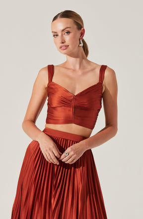 A woman is wearing a rust-colored, ruched crop top with wide shoulder straps and a sweetheart neckline. She is also dressed in a matching high-waisted, pleated skirt made of a shiny fabric.