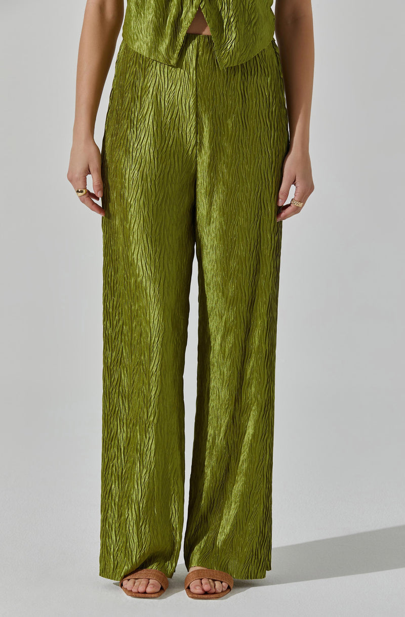 A person is wearing a matching olive green outfit consisting of high-waisted, wide-leg pants with a textured, wavy pattern and a sleeveless top with a similar textured pattern. The top is slightly cropped, showing a small gap at the waist. The person is also wearing open-toe sandals in a light tan color with a block heel, and several rings on their fingers.