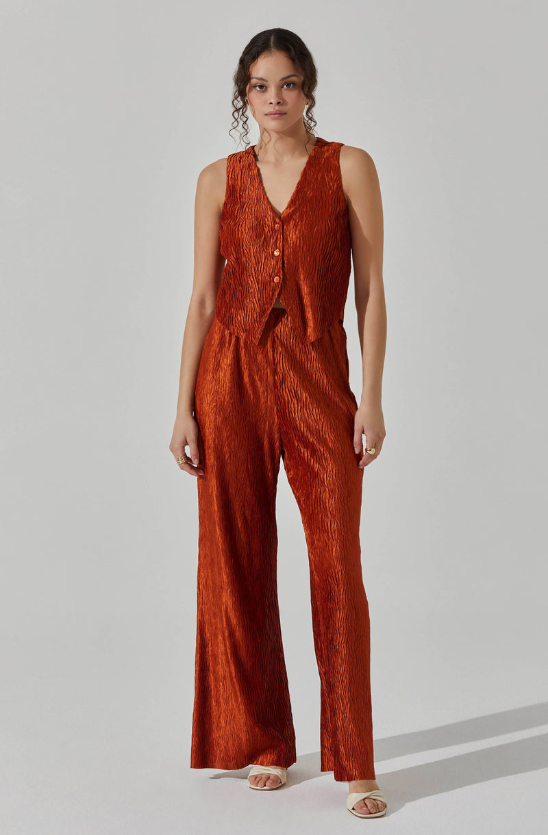 A woman is wearing a sleeveless, V-neck top with a textured, crinkled fabric in a rich, burnt orange color. The top features buttons down the front. She pairs it with matching high-waisted, wide-leg pants made of the same textured fabric.