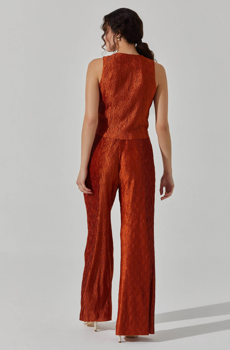 A woman is wearing a sleeveless, burnt orange jumpsuit made of a shiny, textured fabric with subtle vertical striations. The pants are wide-legged and floor-length, and the top has a simple, round neckline.