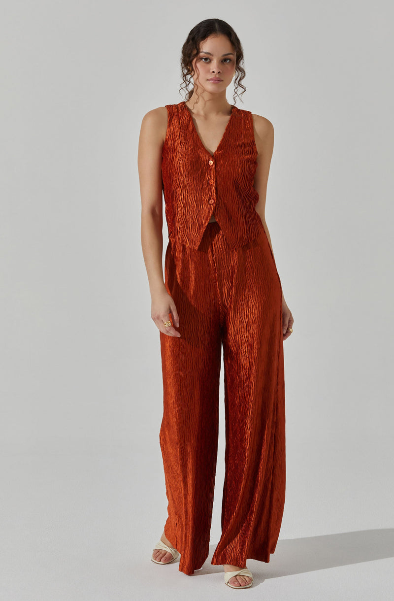 The outfit consists of a sleeveless vest and wide-leg pants, both made from a textured, crinkled fabric in a deep, burnt orange color. The vest features a V-neckline and buttons down the front. The matching pants are high-waisted and flow loosely to the floor. The model is also wearing open-toe shoes in a neutral color.