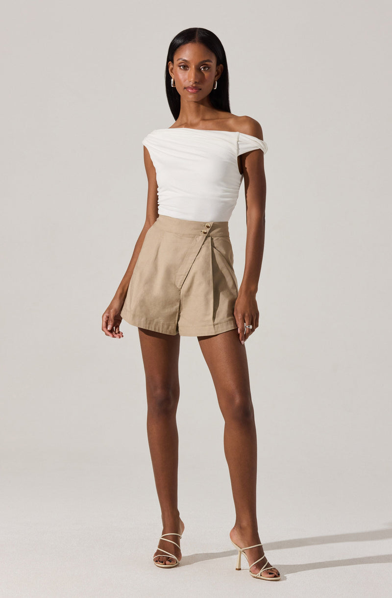 A woman is wearing an off-the-shoulder white top with gathered fabric detailing and short beige high-waisted shorts with a wide waistband and pleats. She is also wearing beige strappy high-heeled sandals.