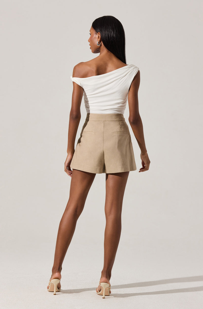 A woman is wearing a white, off-the-shoulder, short-sleeved top with a draped design and beige high-waisted shorts with a fitted waistband and front pockets. She is also wearing beige high-heeled shoes.