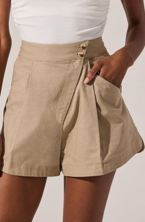 The image shows a person wearing beige high-waisted shorts with pleats at the front and two buttons at the waistband. The shorts have side pockets, and the person has one hand placed inside the pocket. The individual is also wearing a white sleeveless top.