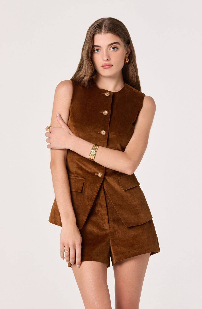 A young woman is wearing a sleeveless, brown suede outfit consisting of a fitted top with an asymmetrical button closure and matching high-waisted shorts with front pockets. She accessorizes with gold hoop earrings, gold rings, and multiple gold bangles on her left wrist.