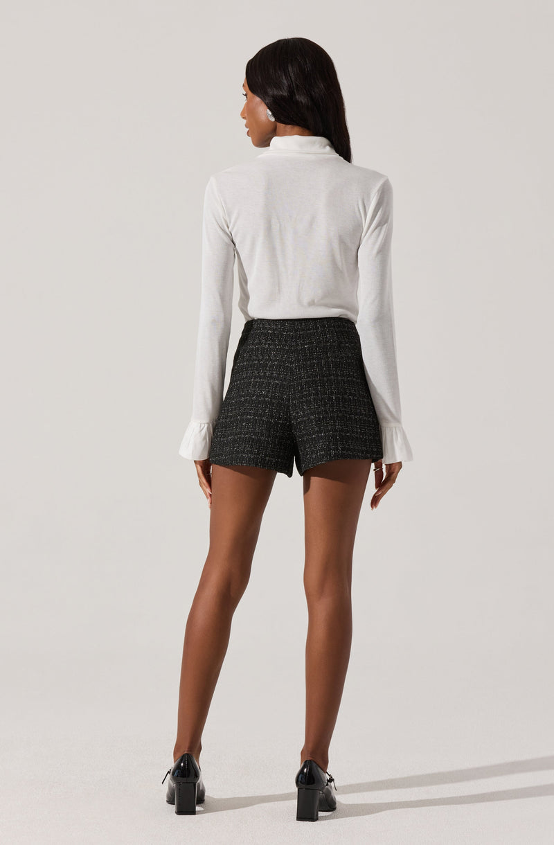 A woman is wearing a white long-sleeve blouse with a high collar and ruffled cuffs, paired with high-waisted black shorts featuring a subtle checkered pattern. She completes the look with black high-heeled shoes.
