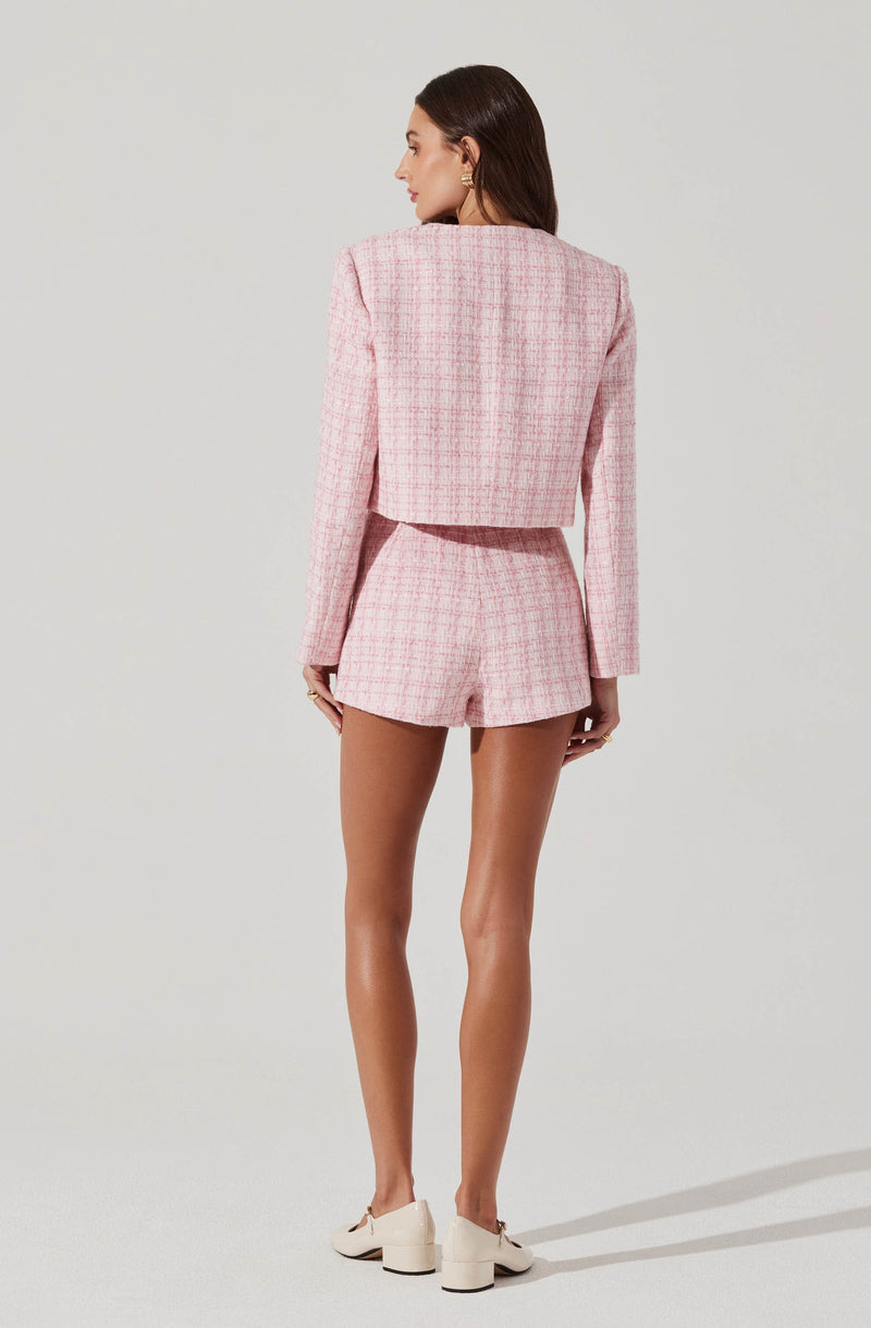 A woman is dressed in a light pink plaid cropped blazer with structured shoulders and long sleeves, paired with matching high-waisted shorts. She is wearing off-white heeled loafers.