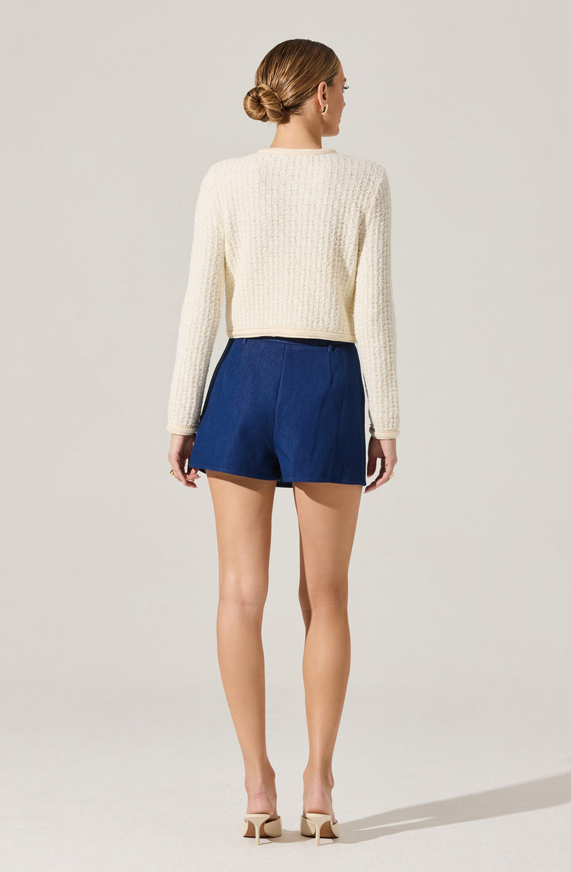 A woman is wearing a cream-colored, chunky knit sweater with long sleeves and a round neckline, paired with high-waisted, dark blue shorts that have a pleated front and a loose fit. She is also wearing nude high-heeled shoes.