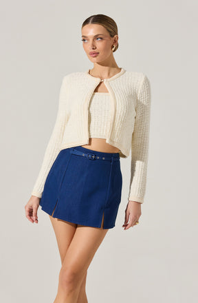 A woman is wearing a textured, cream-colored, cropped cardigan with a round neckline and a front zipper. Underneath, she has a matching cream-colored top. She pairs this with a high-waisted, dark blue denim skirt that features a belt with a small buckle and two front slits.