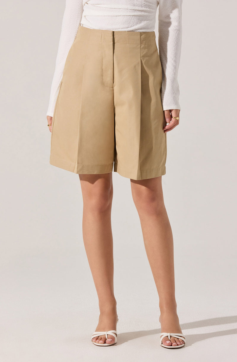 A woman is wearing high-waisted beige shorts with front pleats and wide leg openings, paired with a long-sleeve white top and white open-toe high heels.