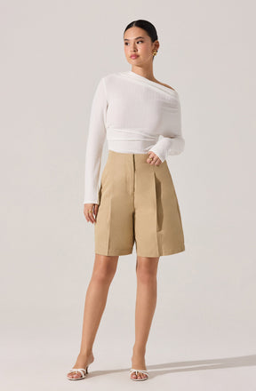 A woman is wearing a white, long-sleeved, asymmetrical sweater with a textured knit pattern, paired with high-waisted, beige knee-length shorts that have a wide-leg fit and pleats at the front. She is also wearing open-toed, strappy heels in a light color.