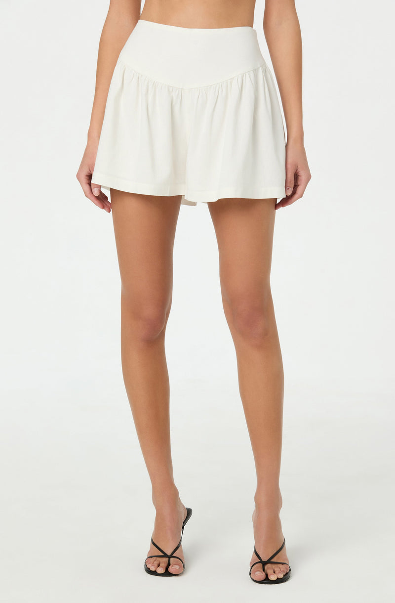 A woman is wearing a high-waisted, white, A-line skort with a gathered or pleated design at the hem. She is also wearing black, strappy high-heeled sandals.