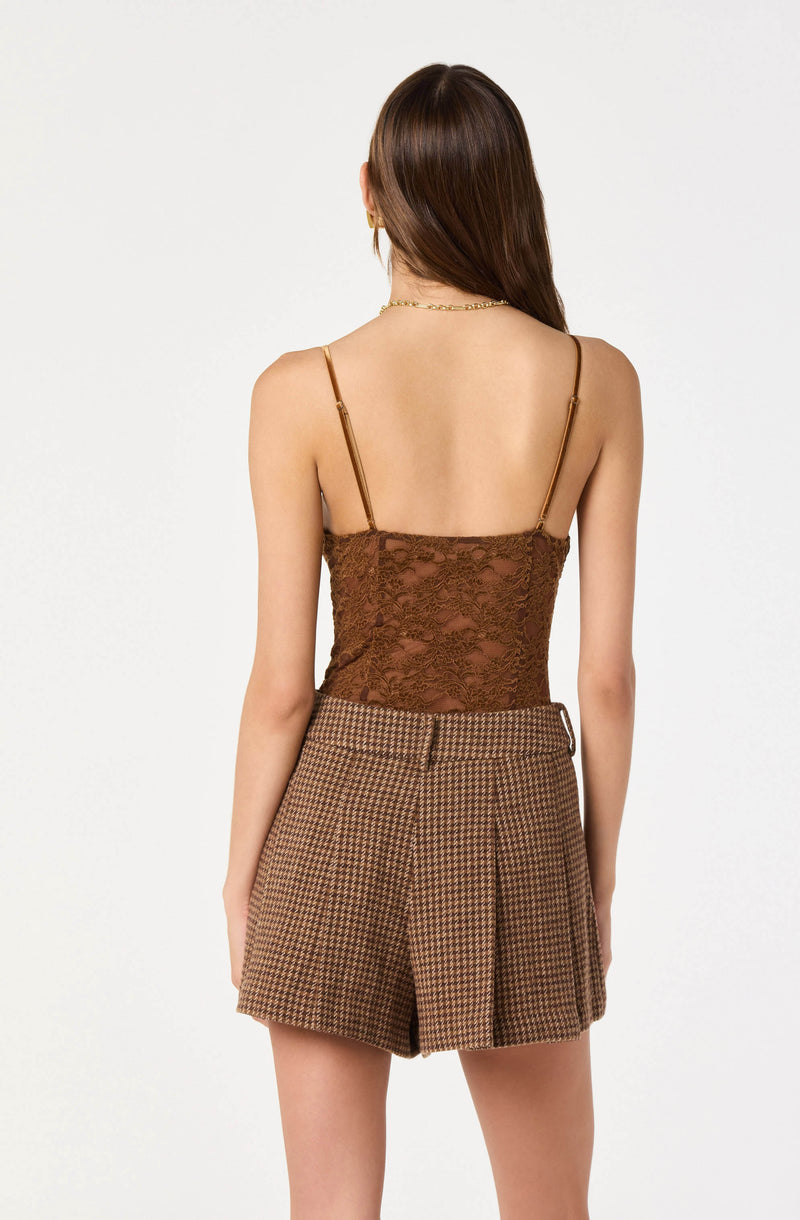 A woman is wearing a brown lace spaghetti strap top with gold adjustable straps. She is also wearing high-waisted shorts with a houndstooth pattern in shades of brown and black.
