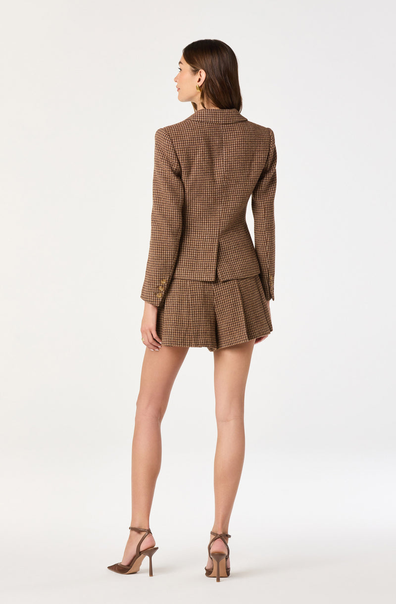A woman is wearing a fitted blazer with a houndstooth pattern in shades of brown, featuring a notched lapel and long sleeves with decorative buttons near the cuffs. She pairs it with matching high-waisted shorts that have pleats, made from the same patterned fabric. She completes the outfit with tan ankle-strap high heels.