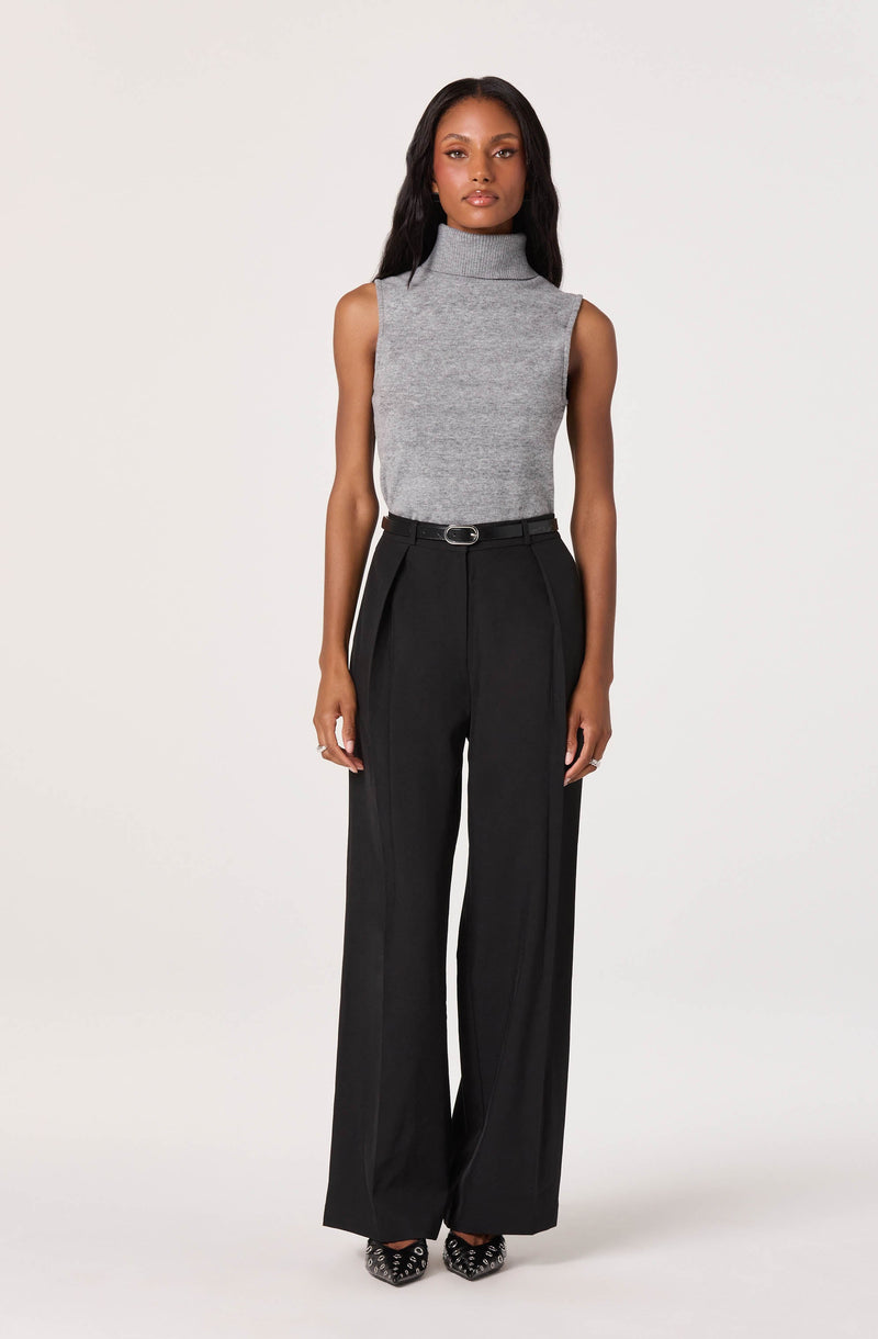 A woman is wearing a sleeveless, high-neck, grey knit top paired with high-waisted, wide-leg black trousers. The trousers have a belt and feature a pleated front design. She completes her outfit with patterned high-heeled shoes.