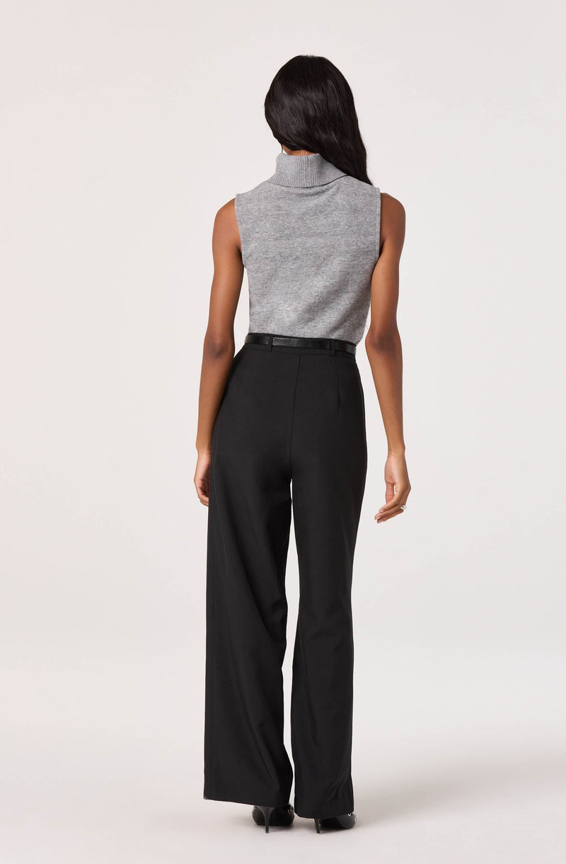 A woman is wearing a sleeveless, gray turtleneck sweater with a high collar that fits snugly around her neck. She is also dressed in high-waisted, black wide-leg trousers that reach the floor, paired with black shoes.