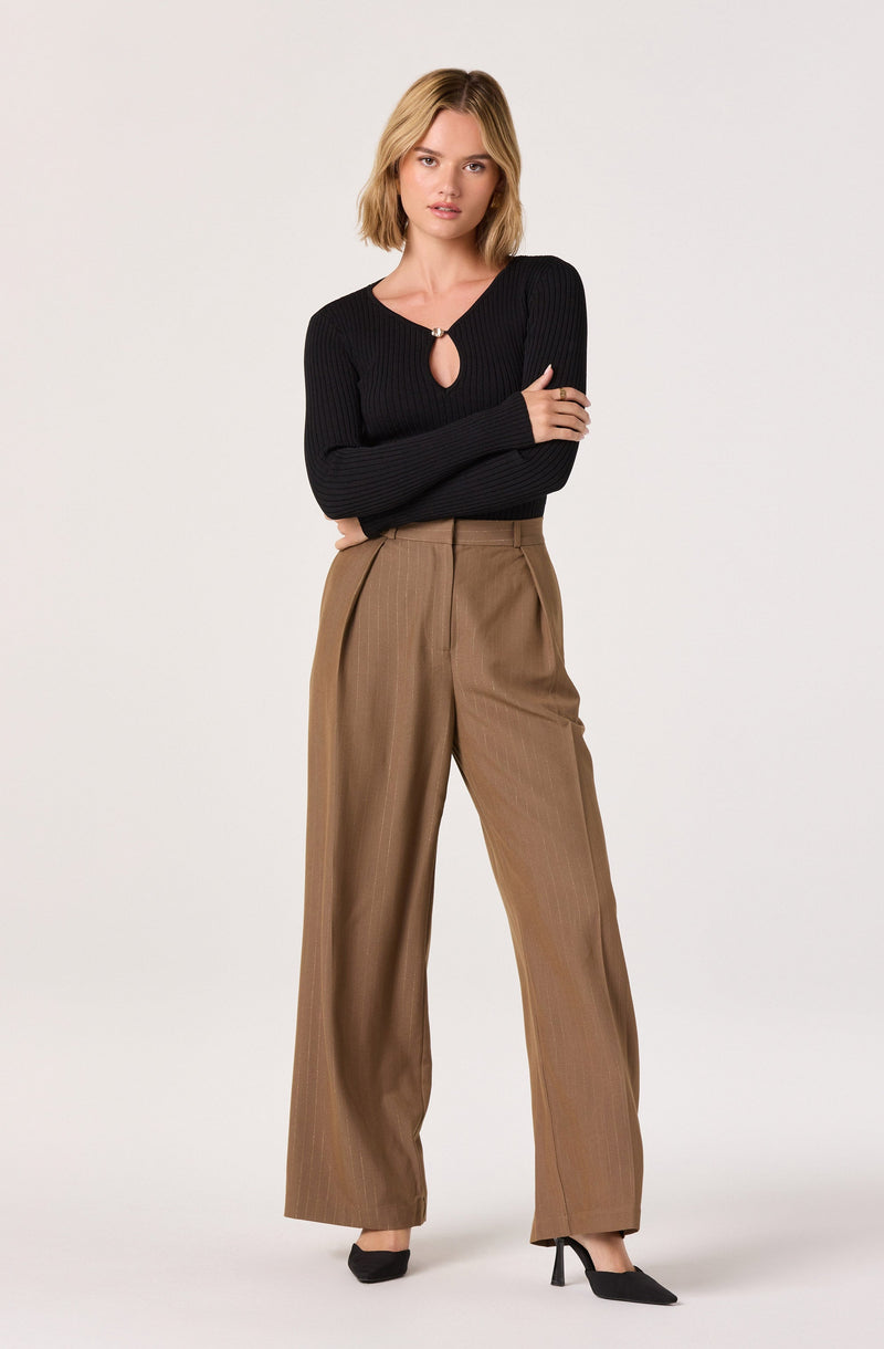 A woman is wearing a fitted, long-sleeved, black ribbed sweater with a small keyhole opening secured by a decorative clasp at the neckline. She is also wearing high-waisted, wide-leg tan trousers with a subtle pinstripe pattern.