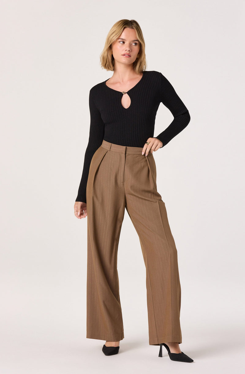 A woman is wearing a fitted black ribbed long-sleeve top with a small keyhole opening at the front, fastened with a metallic ring. She pairs it with high-waisted, wide-leg brown trousers that have pleats at the waist and subtle pinstripes. She completes the outfit with black pointed-toe high heels.