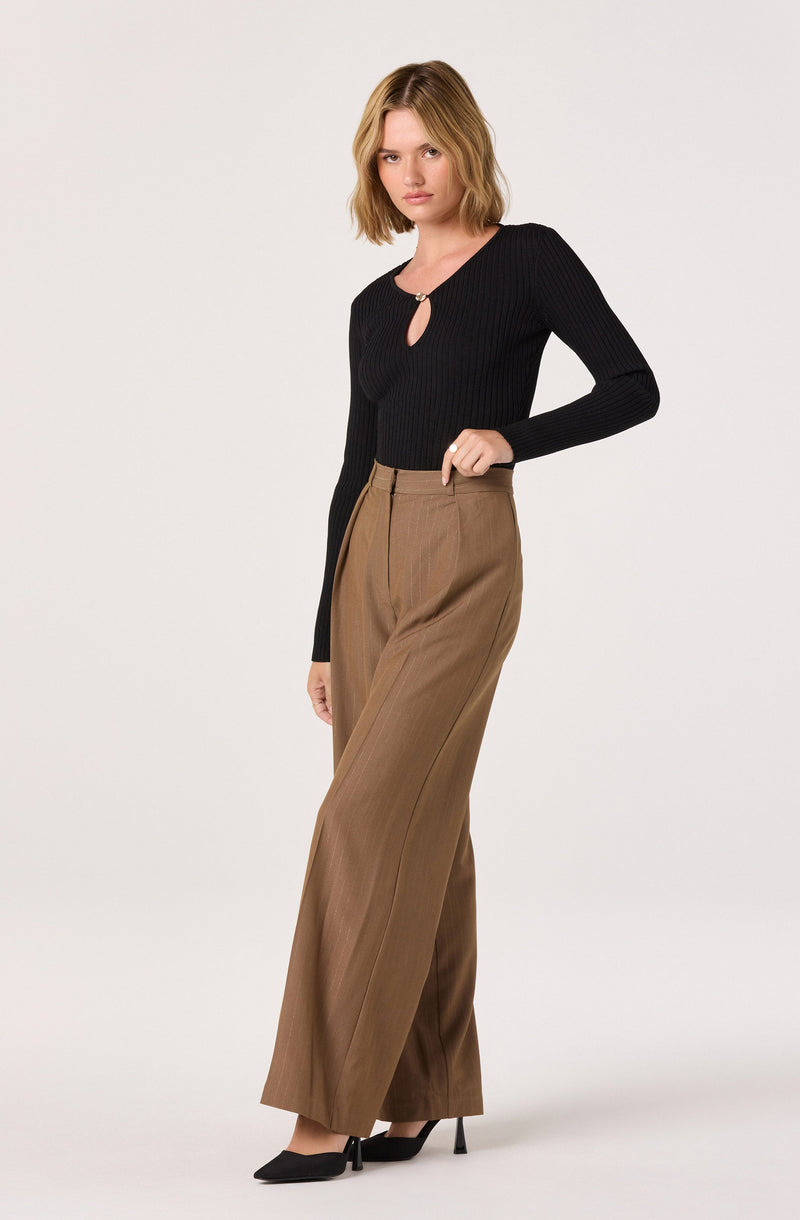 The woman is wearing a black, long-sleeved, ribbed knit top with a small keyhole and metal ring detail at the neckline. She pairs it with high-waisted, wide-leg brown trousers that have a crease down the front. She completes the outfit with black pointed-toe high heels.