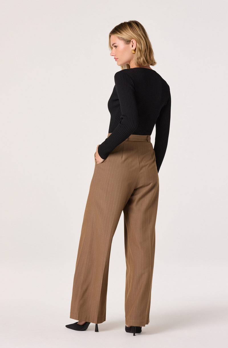 A woman is wearing a fitted black ribbed long-sleeve top with a round neckline, paired with high-waisted, wide-leg brown trousers that have a subtle pinstripe pattern. She is also wearing black pointed-toe high heels.