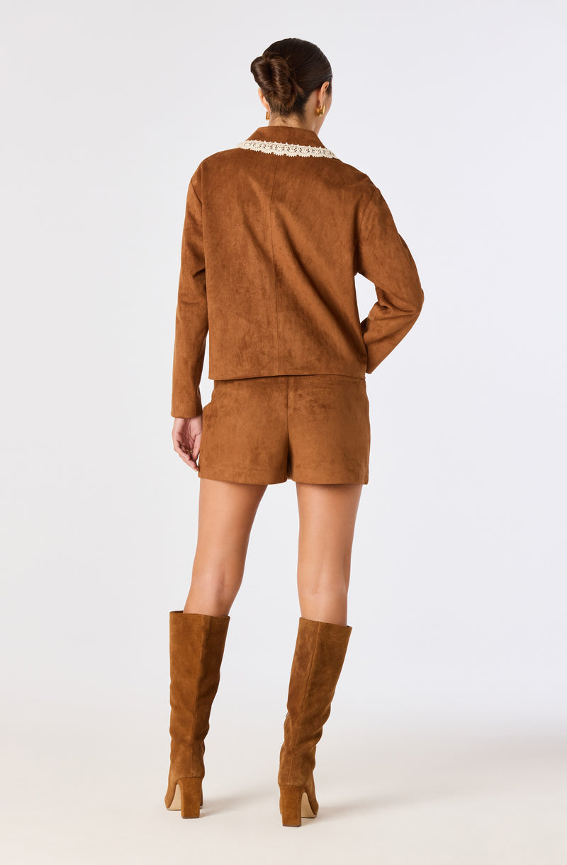 Woman in brown suede jacket, shorts, and knee-high boots, facing away from camera.