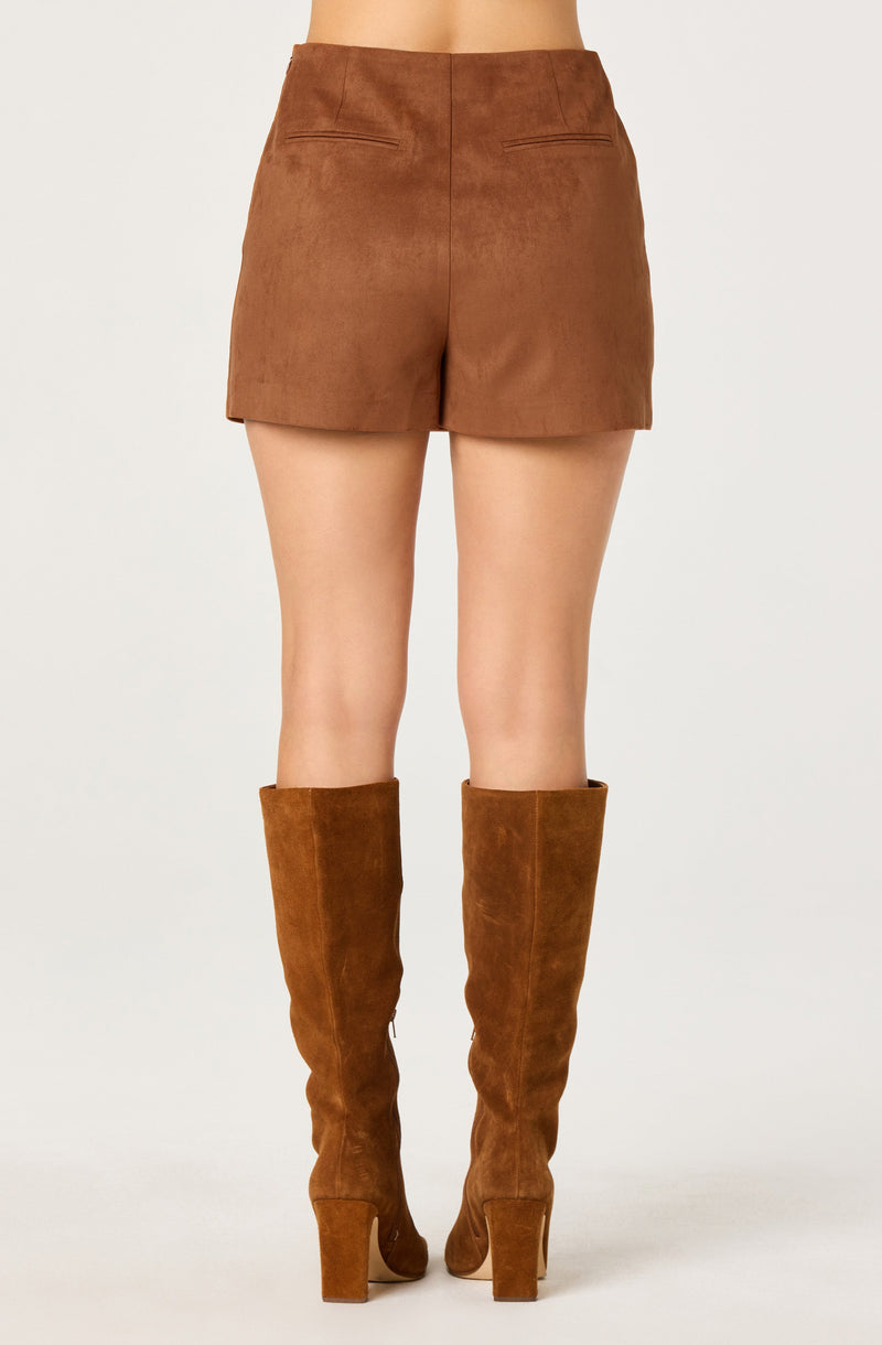 The image shows a person wearing high-waisted, brown shorts made of a soft, suede-like material, with a simple, clean design and two back pockets. The individual is also wearing knee-high, brown suede boots with a chunky heel.