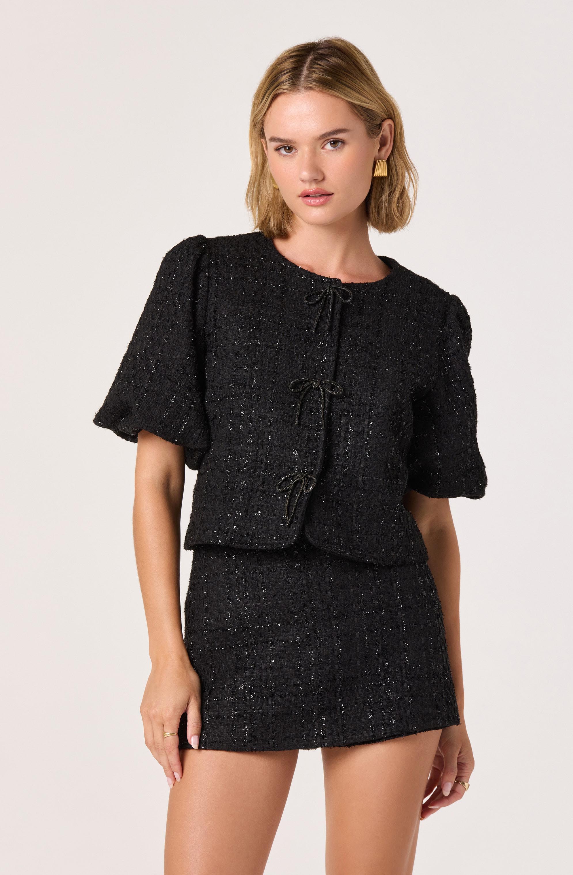 A woman is wearing a black textured two-piece outfit consisting of a short-sleeved, cropped jacket with puffed sleeves and decorative bow ties on the front, paired with a matching high-waisted mini skirt. The fabric has a subtle shimmer and a woven pattern.