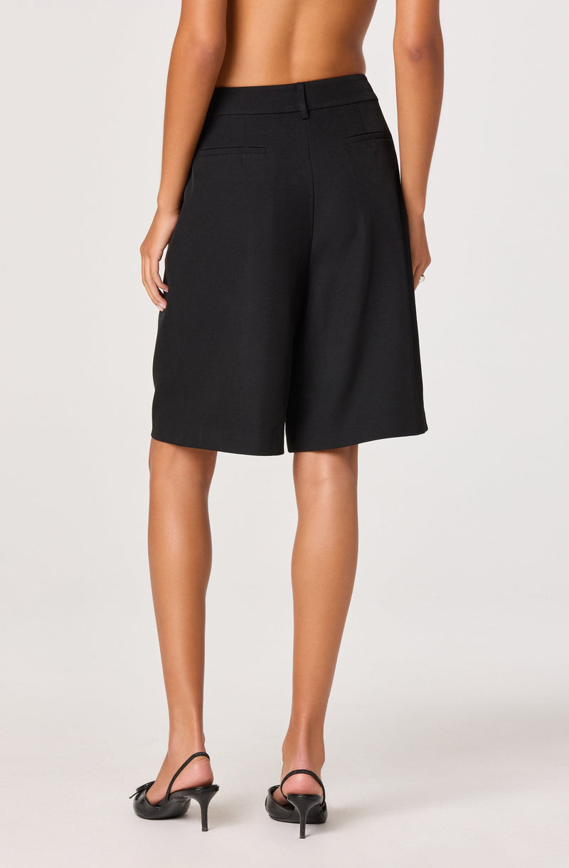 A person is wearing high-waisted, knee-length black shorts with a tailored fit. The shorts have a waistband with belt loops and two horizontal back pockets. The individual is also wearing black high-heeled sandals with thin straps.