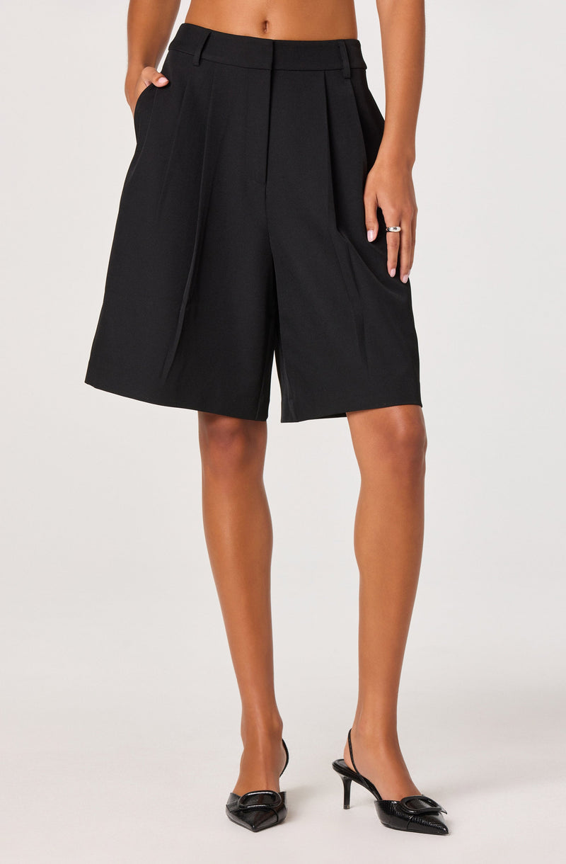 A person is wearing high-waisted black shorts with pleats, front belt loops, and side pockets. The shorts have a loose, wide-leg fit that falls just above the knees. The person is also wearing black pointed-toe high heels with a bow detail on the front and ankle straps.
