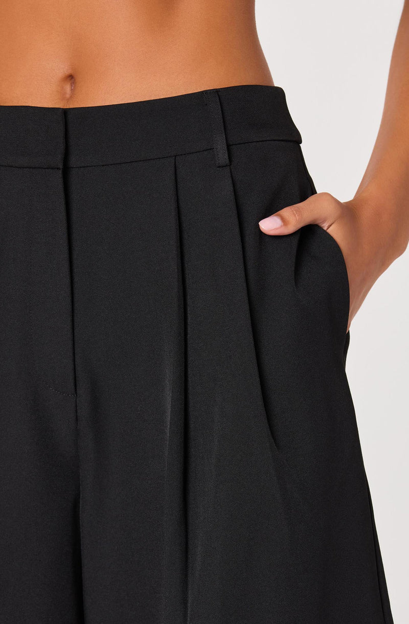 Close-up of a person wearing high-waisted black tailored trousers with a wide waistband, belt loops, and a pleat detail on the front. The person has one hand in the pocket. The fabric appears smooth and structured.